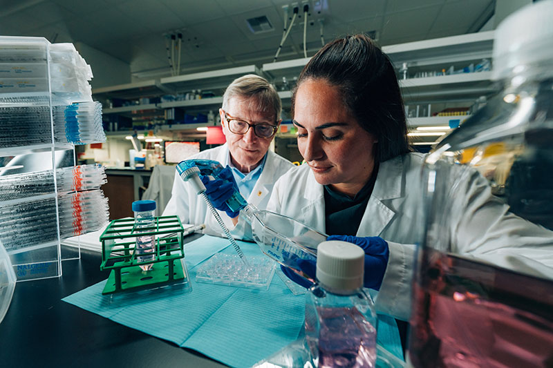 Two scientists in lab coats conduct experiments in a laboratory. One is using a pipette while the other observes.